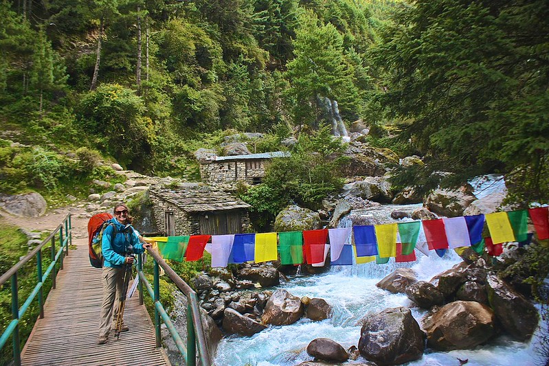 Lina poses by the prayer flags over a creamy blue river. small waterfall in the background