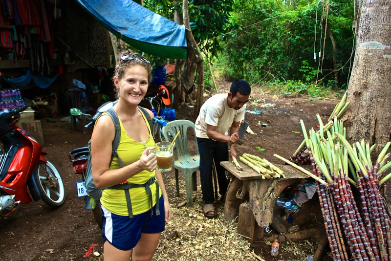 Lina enjoying a drink pressed from raw sugar cane. Man in background, chopping up more sugar cane. Lina enjoying a drink pressed from raw sugar cane. Man in background, chopping up more sugar cane.